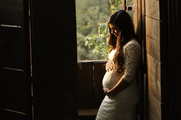 Mother in white lace dress by wooden window in warm light
