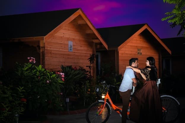 Couple with bicycle by cozy illuminated wooden cabins at night