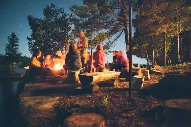 Friends camping around bonfire by lake under starry night